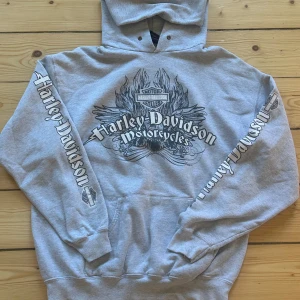 hoodie Harley Davidson y2k 00s rare vintage affliction - Snygg grå hoodie med tryck både framtill och längs ärmarna. Märkt storlek L Mått: Axelbredd 57 cm, längd 66 cm. Tillverkad i UK, 70 % bomull / 30 % polyester. Använd men i gott skick. En mycket liten blå tuschfläck finns på ena ärmen (se bild), knappt synlig vid användning.  y2k, drainer, emo, alt, scene, skater, vintage, grunge, motorcore, harley, biker, oversized, 00s, tumblr, retro, street, early 2000s, flame print, barbed wire, tribal, heavy metal, rare, unisex, collectors item, UK made, USA