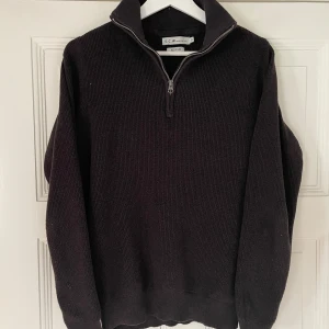 Mörkbrun stickad tröja med dragkedja från S.C.W. - Mysig mörkbrun stickad tröja från S.C.W. med halv dragkedja och hög krage. Tröjan är ribbstickad och passar perfekt för kyligare dagar. Enkel och stilren design som är lätt att matcha.