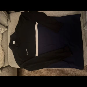 Svart långärmad träningströja från Nike - Säljer en svart långärmad träningströja från Nike med vit logga på bröstet och en vit rand över bröstet. Perfekt för träning eller sport. Tröjan har dragkedja vid halsen och är i ett syntetmaterial.