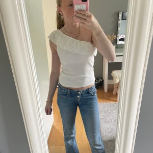 Vit oneshoulder topp med volang - Säljer en vit oneshoulder topp med bred volangkant i spets. Toppen är ribbad och har en snygg One shoulder. Perfekt för dig som vill ha en unik och trendig look. Passar perfekt i sommar speciellt med ett par jeans eller jeans kjol.