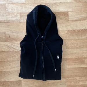 Svart hoodie från Polo Ralph Lauren - Säljer en svart hoodie från Polo Ralph Lauren med dragkedja och vit broderad logga på bröstet. Tröjan har huva med snörning och två fickor framtill. Perfekt för en avslappnad stil.