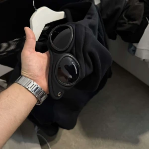 Svart hoodiejacka med goggles från C.P. Company - Svart hoodiejacka från C.P. Company med ikoniska goggles i huvan. Jackan har dragkedja framtill, två fickor med dragkedja och justerbar huva med snörning. Perfekt för dig som gillar streetwear och unika detaljer.