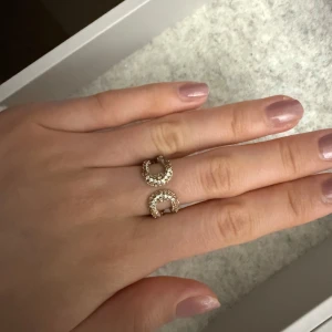 Guldfärgad ring med dubbla cirklar och stenar - Trendig guldfärgad ring med två öppna cirklar dekorerade med små glittrande stenar. Snygg och unik design som sticker ut på handen. Går att justera efter din egna storlek 