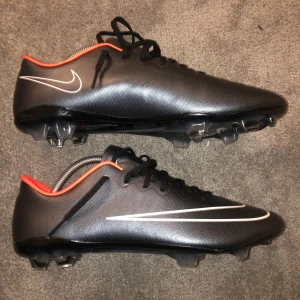 Nike Mercurial fotbollskor - Säljer ett par svart/gråa Nike fotbollsskor med vit swoosh och röda detaljer vid öppningen. Skorna har snörning och är designade för bra grepp på planen. ACC (All Conditions Control) teknologi för optimal bollkontroll i alla väder.