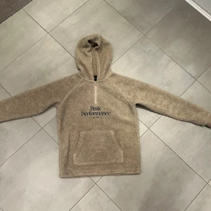 Beige fleece hoodie från Peak Performance - Mysig beige hoodie från Peak Performance i fleece-material. Tröjan har huva, halv dragkedja framtill och en stor magficka. Perfekt för kyliga dagar och enkel att matcha med andra plagg.