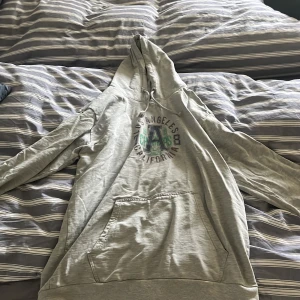 Grå hoodie från Lager 157 med Los Angeles-tryck - Säljer en grå hoodie från Lager 157 i storlek XL. Tröjan har tryck med texten 'Los Angeles California Champs' och stora bokstaven A på bröstet. Klassisk känguruficka framtill och huva. Perfekt för en avslappnad stil.