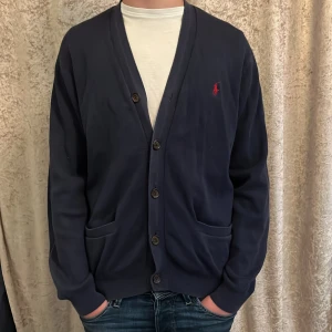 Mörkblå kofta från Polo Ralph Lauren - Mörkblå kofta från Polo Ralph Lauren med knäppning framtill och två fickor. Klassisk design med röd broderad logga på bröstet. Perfekt för sommarkvällar. Skick är 9/10 och nypris är 2799 kr!!