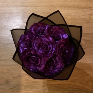 Lila bukett med satinblommor - Bukett med stora lila rosor i satinliknande material, arrangerade som en bukett och insvepta i svart. Perfekt present