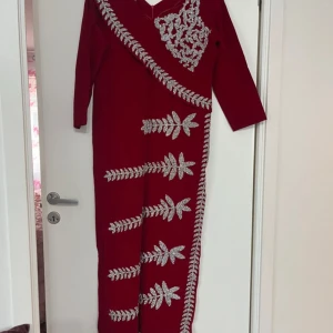 Vinröd långklänning med silverbroderi - Säljer en vinröd långklänning med långa ärmar och v-ringning. Klänningen har vackra silverbroderade bladmönster och detaljer längs hela framsidan och axeln. Perfekt för festliga tillfällen när du vill sticka ut. Obs: referens bild 