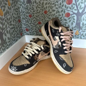 Nike SB Dunk Low Travis Scott - Nike SB Dunk Low Travis Scott med beige och svart färgskala, paisley- och rutigt mönster samt olika färger på skosnörena. Skorna har rund tå och platt sula, och är tillverkade i skinn och textil. Säljs med originalkartong. Tyvärr ej kvitto var ett bra tag sen de var köpta. Billigt pris på grund av att jag ej har kvitto kvar och ganska gamla. Pris kan förhandlas
