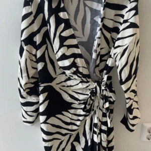 Omlottklänning med knytband - Snygg omlottklänning i svart och vitt med knytband i midjan. Klänningen har lång ärm med split. Köpt på H&M. Storlek M. Använd ytterst få gånger, i väldigt fint skick.