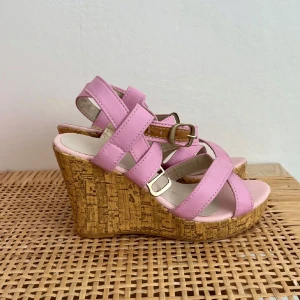 Rosa kilklack sandaler  - Säljer ett par snygga sandaler med rosa remmar och justerbart spänne. Skorna har en hög kilklack i kork och öppen tå, perfekt för varma dagar. Ger både höjd och komfort med sin stabila plattform.
