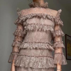 Beige spetsklänning med volanger från By Malina - Supersöt beige spetsklänning från By Malina med massor av volanger och hög krage. Klänningen har trekvartsärm och är täckt av fina spetsdetaljer. Perfekt för dig som vill sticka ut med en romantisk look✨💕