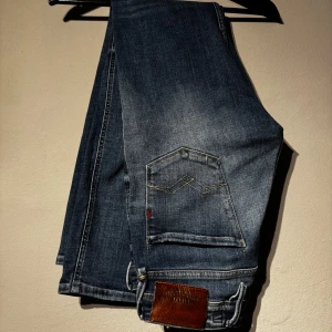 Replay jeans - Säljer ett par blå jeansbyxor från Replay med slitningar och lätt tvättad look. Byxorna har klassisk femficksdesign, raka ben och läderpatch bak i midjan. Perfekta för en avslappnad stil.