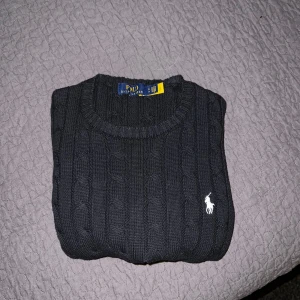 Svart stickad tröja, Polo Ralph Lauren - Säljer en klassisk svart stickad tröja från Polo Ralph Lauren med rund halsringning och vit broderad logga på bröstet. Tröjan har kabelstickat mönster och ribbade muddar vid ärmslut och nederkant. Perfekt för en stilren look. Bra för storlek XS, mdn funkar också perfekt för S om man vill ha den ganska tight för att visa sin kroppsbyggnad.