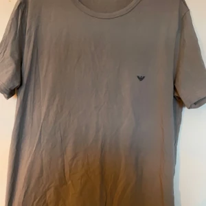 Grå t-shirt från Emporio Armani - Säljer en stilren grå t-shirt från Emporio Armani med liten logga på bröstet. T-shirten har rund halsringning och är tillverkad i mjuk bomullsblandning. Perfekt för en clean och enkel look.