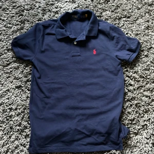 Mörkblå pikétröja från Polo Ralph Lauren - Säljer en mörkblå pikétröja från Polo Ralph Lauren med röd broderad logga på bröstet. Tröjan har korta ärmar, klassisk krage och två knappar framtill. Slim fit-modell som sitter snyggt och stilrent. Tröjan är använd MAX 5 gånger och då bara lugit i min garderob. 