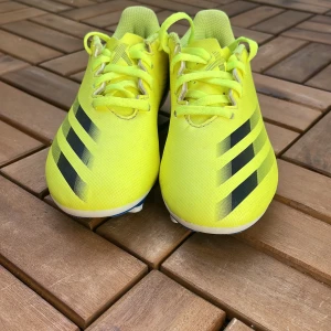 Gula fotbollsskor från Adidas - Säljer ett par riktigt snygga gula fotbollsskor från Adidas med svarta ränder på sidorna. Skorna har snörning och är tillverkade i syntetmaterial. Perfekta för dig som vill sticka ut på planen!