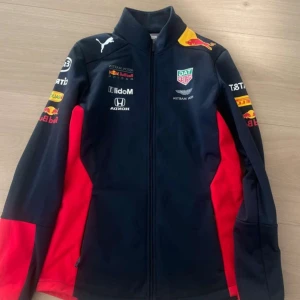 Red Bull Racing jacka med loggor - Sportig jacka från Red Bull Racing med flera tryckta loggor, dragkedja framtill och inslag av rött och gult på marinblå bas. Jackan har lång ärm och hög krage. 