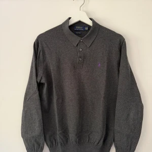 Grå långärmad pikétröja från Polo Ralph Lauren - Säljer en klassisk grå pikétröja från Polo Ralph Lauren i mjuk pima-bomull. Tröjan har lång ärm, krage och knäppning med tre knappar framtill. Liten broderad logga på bröstet i lila. Perfekt för en stilren look.
