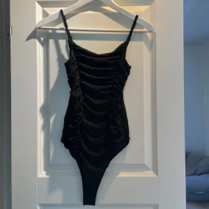 Svart croppad bodysuit med rynkade detaljer - Säljer en svart bodysuit med smala axelband och rynkade detaljer framtill.