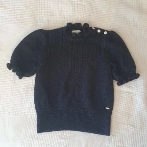 Svart glittrig stickad tröja från Holly & Whyte - Supersöt svart stickad tröja med glittriga trådar från Holly & Whyte. Tröjan har puffärmar, volangdetaljer vid hals och ärmslut samt tre blommiga knappar på axeln. Perfekt för dig som vill ha något extra i vardagen!💘