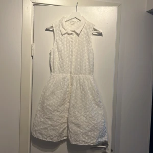 Vit ärmlös klänning med krage och blommigt broderi - Säljer en söt vit ärmlös klänning med klassisk krage och knäppning framtill. Klänningen har ett broderat blommönster och är tillverkad i ett lätt tyg med fodrad insida. Perfekt för sommarens tillfällen och studentfirande! Är ej genomskinlig och mycket bra i skicket 