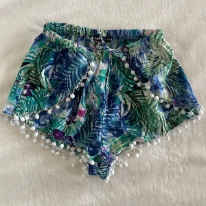Blommiga shorts med tofsar från Bik Bok - Färgglada shorts från Bik Bok med tropiskt blommönster i grönt, blått och lila. Shortsen har vita tofsar längs kanterna och elastisk midja med knytband. Perfekta för varma dagar och strandhäng.