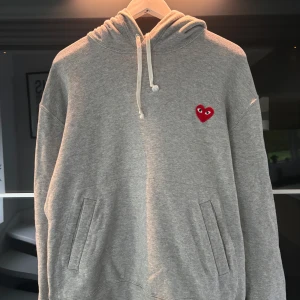 Grå hoodie från Comme des Garçons Play - Säljer en grå hoodie från Comme des Garçons Play med det ikoniska röda hjärtat broderat på bröstet. Tröjan har huva med dragsko och fickor framtill. Perfekt för en avslappnad och stilren look. Storlek l men passar m