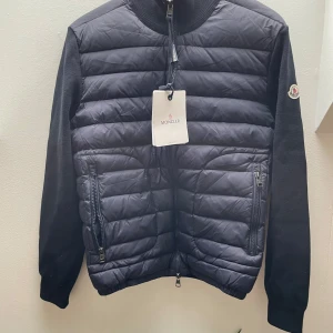 Mörkblå cardigan från Moncler - Säljer en mörkblå cardigan från Moncler med stickade ärmar och quiltad framsida. Jackan har dragkedja framtill och två fickor med dragkedja. Klassisk Moncler-logga på ärmen och hög krage.