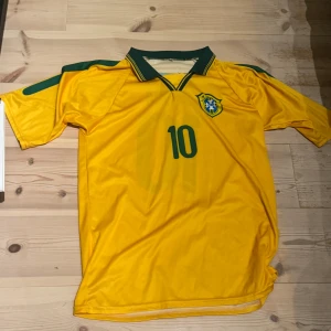 Brasilien fotbollströja Kaká #10 - Gul Brasilien fotbollströja med gröna detaljer, nummer 10 och Kaká på ryggen. Klassisk design med grönt krage och landslagets emblem på bröstet. Perfekt för dig som älskar fotboll. Storlek M och är endast använd 1 gång.