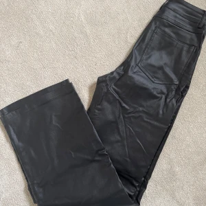 Svarta byxor i skinnimitation - Snygga svarta byxor i skinnimitation med hög midja och raka, vida ben. Klassisk femficksmodell med bälteshällor och stilren look. Perfekta för att lyfta vilken outfit som helst. Ej använda! 