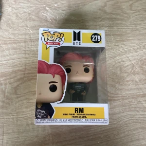 Funko Pop! Rocks BTS RM figur - Säljer en Funko Pop! Rocks vinylfigur av RM från BTS, nummer 279. Figuren har rosa hår och svarta ögon, och kommer i originalförpackning med BTS-logga. Perfekt för samlare eller fans av K-pop och BTS.
