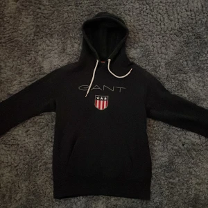Svart hoodie från GANT med logga - Säljer en svart hoodie från GANT i storlek M. Tröjan har vit dragsko i huvan och en broderad GANT-logga med sköld och stjärnor på bröstet. Klassisk och stilren modell med lång ärm och mjukt material.