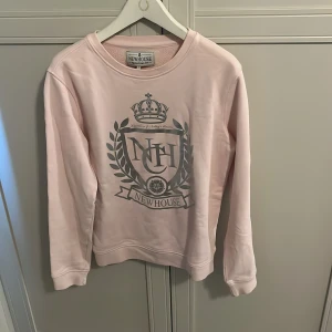 Ljusrosa sweatshirt från Newhouse - Säljer en ljusrosa sweatshirt från Newhouse med stort tryck framtill i grått med krona och emblem. Tröjan har rund halsringning och långa ärmar. Perfekt för en avslappnad och stilren look.