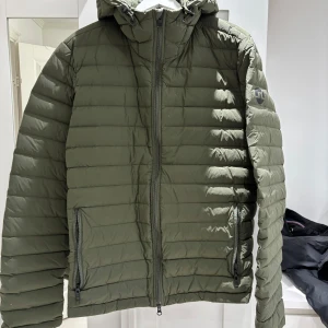 Jacka Peak Performance - Säljer en olivgrön pufferjacka från Peak Performance med huva och dragkedja framtill. Jackan har två sidofickor med dragkedja och diskret logga på ärmen. Perfekt för kyliga dagar.