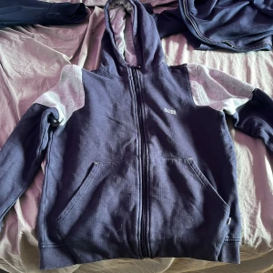 Mörkblå hoodie med dragkedja från Hugo Boss - Säljer en mörkblå hoodie från Hugo Boss med dragkedja och huva. Jackan har vita och grå detaljer på axlarna och ärmarna samt två fickor framtill. Perfekt för en avslappnad stil.