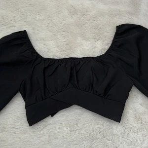 Svart croppad blus med rosett - Trendig svart croppad blus med vida långa ärmar och stor rosett baktill. Blusen har en lätt rynkad byst och bred resår vid axlarna för en snygg passform. 