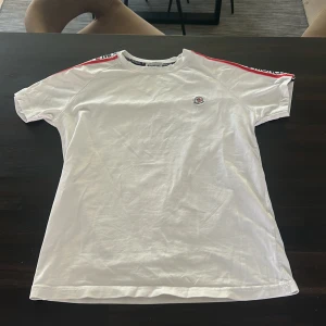 Vit t-shirt från moncler - Vit t-shirt från Moncler med korta ärmar och röd/blå band längs axlarna där Moncler-loggan syns. Har kvitta på den så kan skicka med det om man vill!
