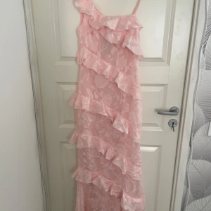 Ljusrosa långklänning med volanger - Säljer en ljusrosa långklänning med volanger och ett axelband. Klänningen har blommigt mönster i tyget och asymmetrisk volangdetalj över hela klänningen. Från boohoo och ny med prislapp.