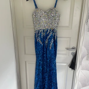 Blå långklänning med paljetter och stenar - Säljer en glittrig blå långklänning med smala axelband. Överdelen är täckt av silvriga paljetter och stora stenar, medan nederdelen är helt blå med paljetter. Perfekt för bal eller fest. Figurnära passform och hjärtformad urringning. Köpt för längesedan så vet ej vart den är ifrån