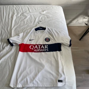 PSG hemmatröja 2023/24 - Vit Paris Saint-Germain fotbollströja med blått och rött band över bröstet samt Qatar Airways sponsortryck. PSG-emblem och Nike-logga på bröstet. Klassisk rund hals och korta ärmar. Perfekt för fans av PSG. Väldigt fin och oanvänd tröja. Den är använd 1-2 gånger ungefär.  Helt ny från Zalando där ny priset ligger runt 1200. 