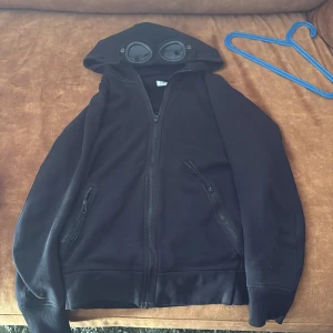 Svart hoodie med glasögon från C.P. Company - Svart hoodie från C.P. Company med ikoniska glasögon i huvan. Jackan har dragkedja framtill, två fickor med dragkedja och ribbade muddar. Perfekt för dig som gillar streetwear och unika detaljer. Ett litet hål syns på bild 5.