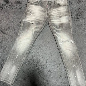  DSQUARED2 jeans - Säljer ett par grå jeans från DSQUARED2 med slitningar och färgstänk för en cool, använd look. Jeansen har raka ben, klassisk femficksdesign och dragkedja. Perfekta för dig som gillar streetstyle och vill sticka ut. storlek 34 Äkta!!!