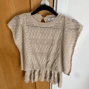 Beige stickad croppad topp från Zara, stl S - Säljer en beige stickad croppad topp från Zara med korta ärmar och volangdetalj nedtill, använd endast ett par gånger, jättefint skick! Toppen har ett fint hålmönster och knapp i nacken. Perfekt för att lyfta din outfit!