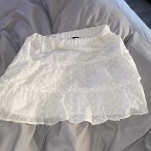 Vit volangkjol med broderade detaljer - Säljer en söt vit kortkjol med volanger och broderade blommönster. Kjolen har resår i midjan och är perfekt för varma dagar. Lätt och luftig känsla med fina detaljer.