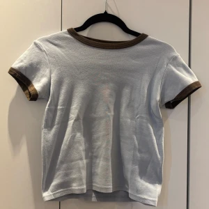 Vit t-shirt med bruna detaljer från Brandy Melville - Vit t-shirt från Brandy Melville med bruna muddar vid halsringning och ärmslut. Klassisk passform och tillverkad i 100% bomull. Perfekt basic-plagg med retrovibbar. Passar XS-S