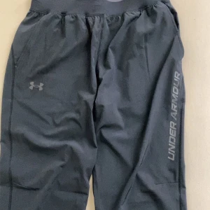 Svarta träningsbyxor från Under Armour - Säljer ett par svarta träningsbyxor från Under Armour med resår i midjan och logga på benet. Byxorna har rak passform och är perfekta för träning eller sport. Materialet är lätt och syntetiskt för bra rörelsefrihet. Inte använda alls, köpte fel storlek