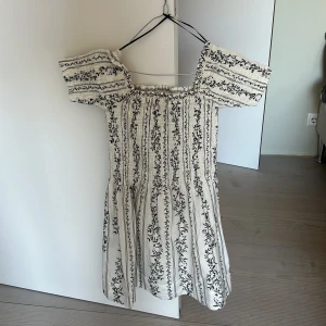 Vit blommig klänning med puffärmar - Supersöt vit klänning med svarta blommiga mönster och puffiga korta ärmar. Klänningen har smock upptill och en luftig kjol, perfekt för varma dagar. Den har en romantisk och trendig känsla med fina detaljer.