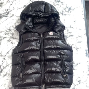Svart dunväst med huva från Moncler - Svart Moncler dunväst i riktigt bra skick med glansig finish och avtagbar huva. Västen har två fickor med dragkedja och klassisk Moncler-logga på bröstet och med NFC scan. Storlek S men kan eventuellt passa M. Vid frågor eller funderingar skicka ett pm, Mvh